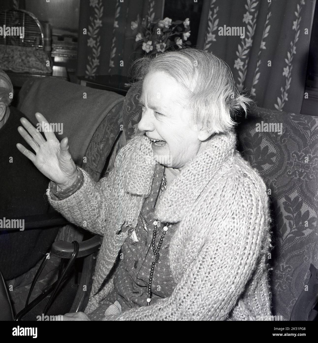 1964, historisch, eine lebhafte ältere Dame, die einen großen Baumwollcardigan trägt, lacht auf ihrem Stuhl in einem Altersheim. Stockfoto