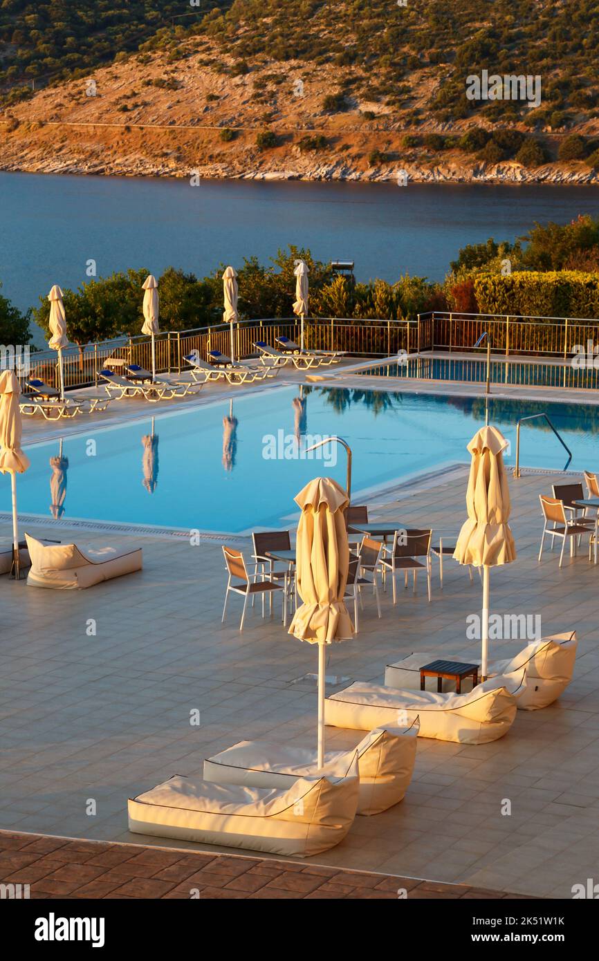 Luxuriöser Swimmingpool am Morgen mit leeren Liegestühlen, Tischen und Sonnenschirmen im Resort mit wunderschönem Meerblick. Griechische Insel Stockfoto