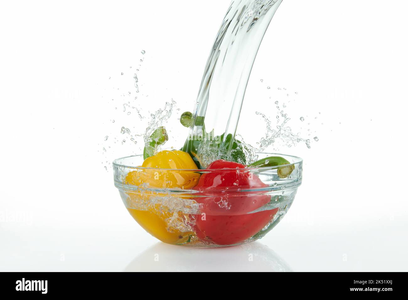 Wasserfall, der auf eine Glasschüssel mit bunten Paprika mit Spritzern auf weißem Hintergrund gießt. Stockfoto