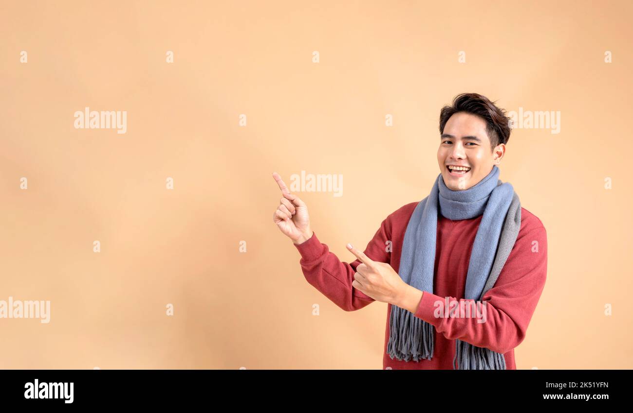 Positiver asiatischer Mann in rotem Pullover und Schal, der mit den Fingern auf einen leeren Bereich zeigt, der auf beigefarbenem Hintergrund isoliert ist. Fröhliche modische 20s Mann beiseite zeigen Geste - halbe Körperlänge Studio-Aufnahme. Stockfoto