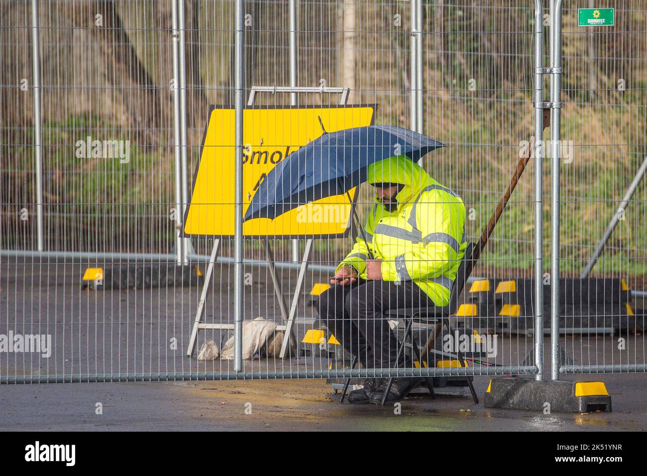 Sicherheitsmann, in gelber Jacke, sitzt unter einem Regenschirm hinter Metallzäunen, die im Dienst an einer Covid-19-Teststation im Regen stehen, Dezember 2020. Stockfoto