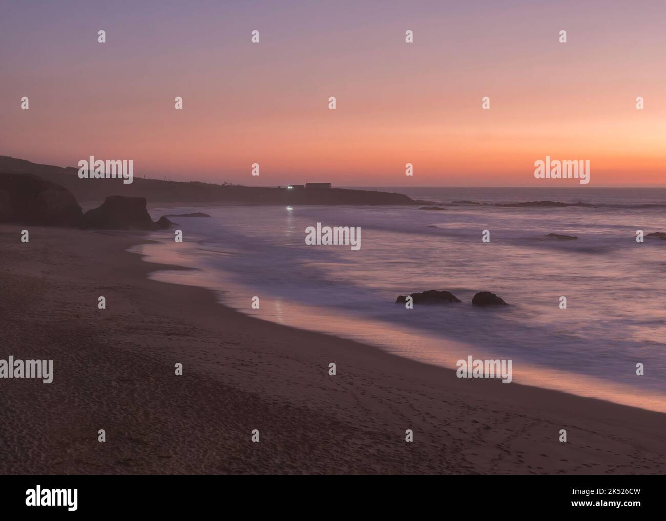 Geheimnisvolle Langzeitansicht des Sandstrandes Praia Grande de Almograve mit verschwommenen Wellen in rosa orange und violettem Licht. Blaue Stunde nach Sonnenuntergang Stockfoto