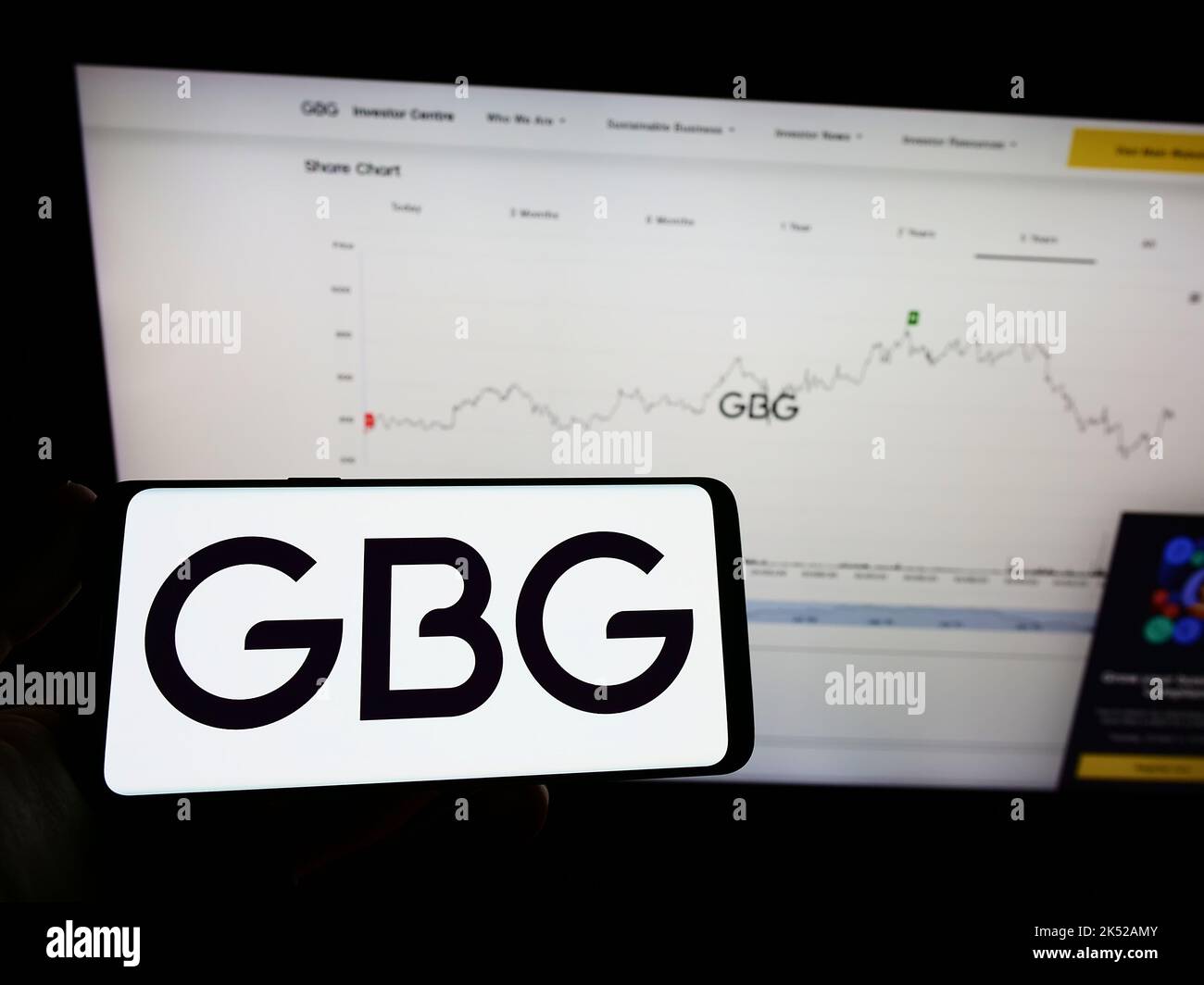 Person mit Mobiltelefon und Logo der Firma GB Group plc (GBG) auf dem Bildschirm vor der Webseite. Konzentrieren Sie sich auf die Telefonanzeige. Stockfoto