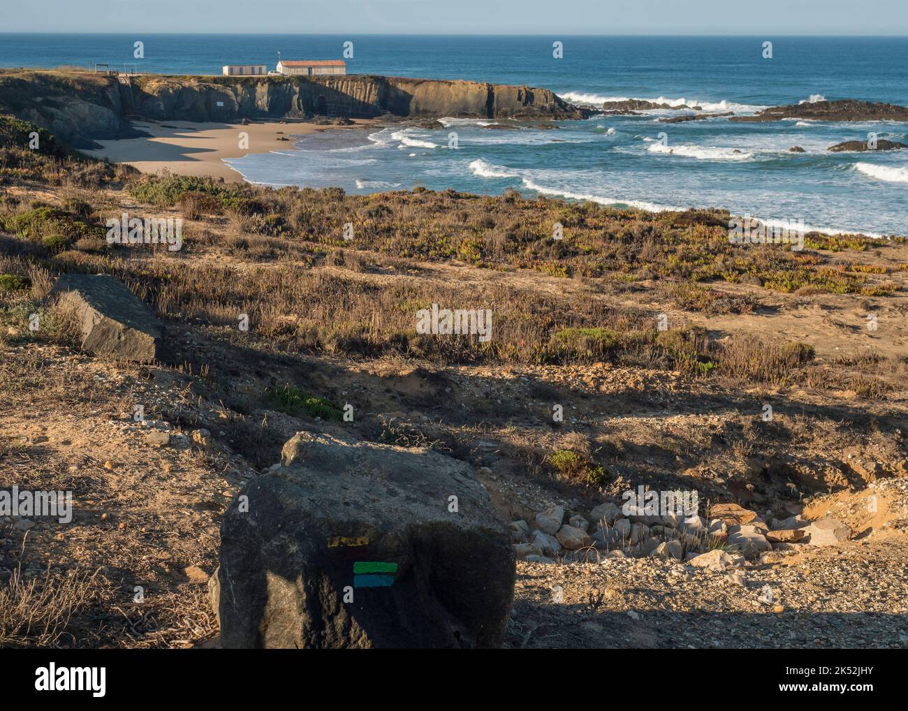 Stein mit blauem und grünem Zeichen des Fischerwegs und Blick auf den Praia Grande de Almograve Sandstrand mit Meereswellen und kleines Strandhaus. Klares Blau Stockfoto