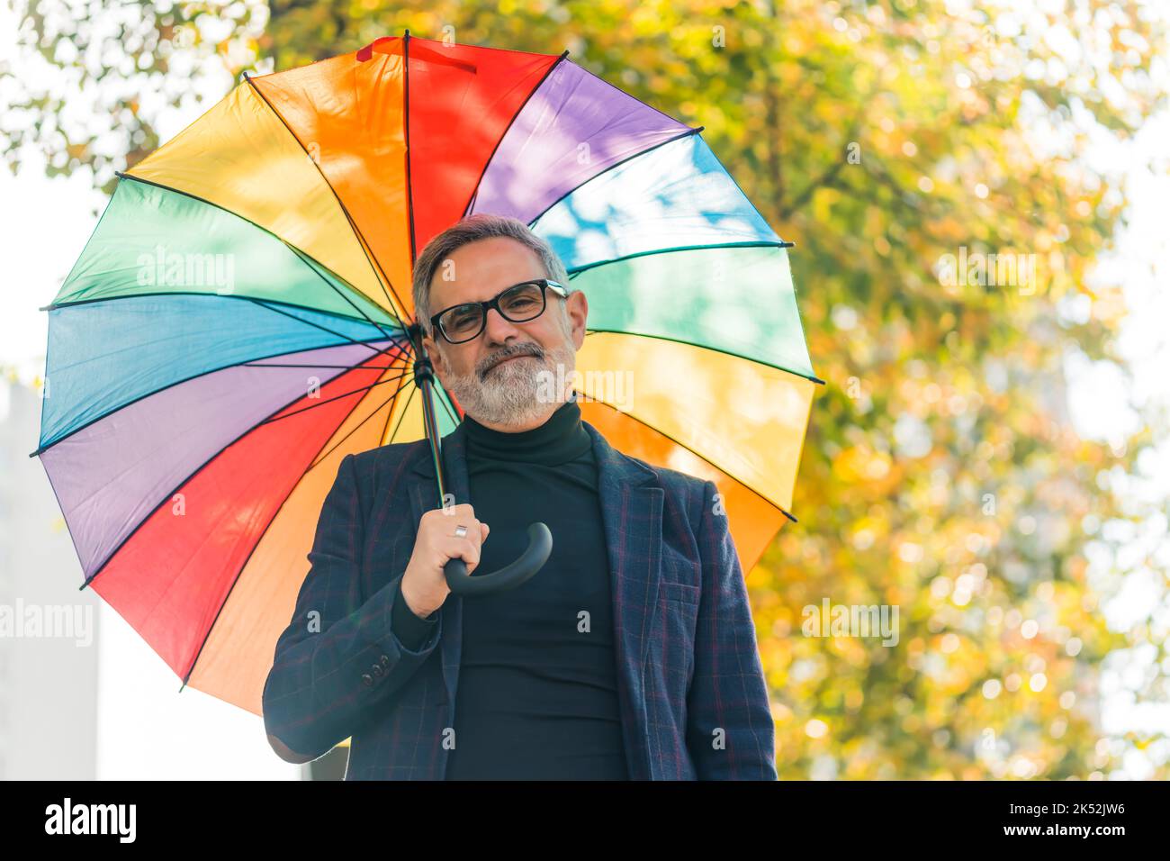 Schönes Outdoor-Porträt von positiven und lebensvollen älteren kaukasischen grauhaarigen Mann in Blazer und Rollkragen Blick auf die Kamera, während er seinen Kopf mit lebendigen Regenbogenschirm. Unscharfe Bäume mit gelben Blättern im Hintergrund. Hochwertige Fotos Stockfoto