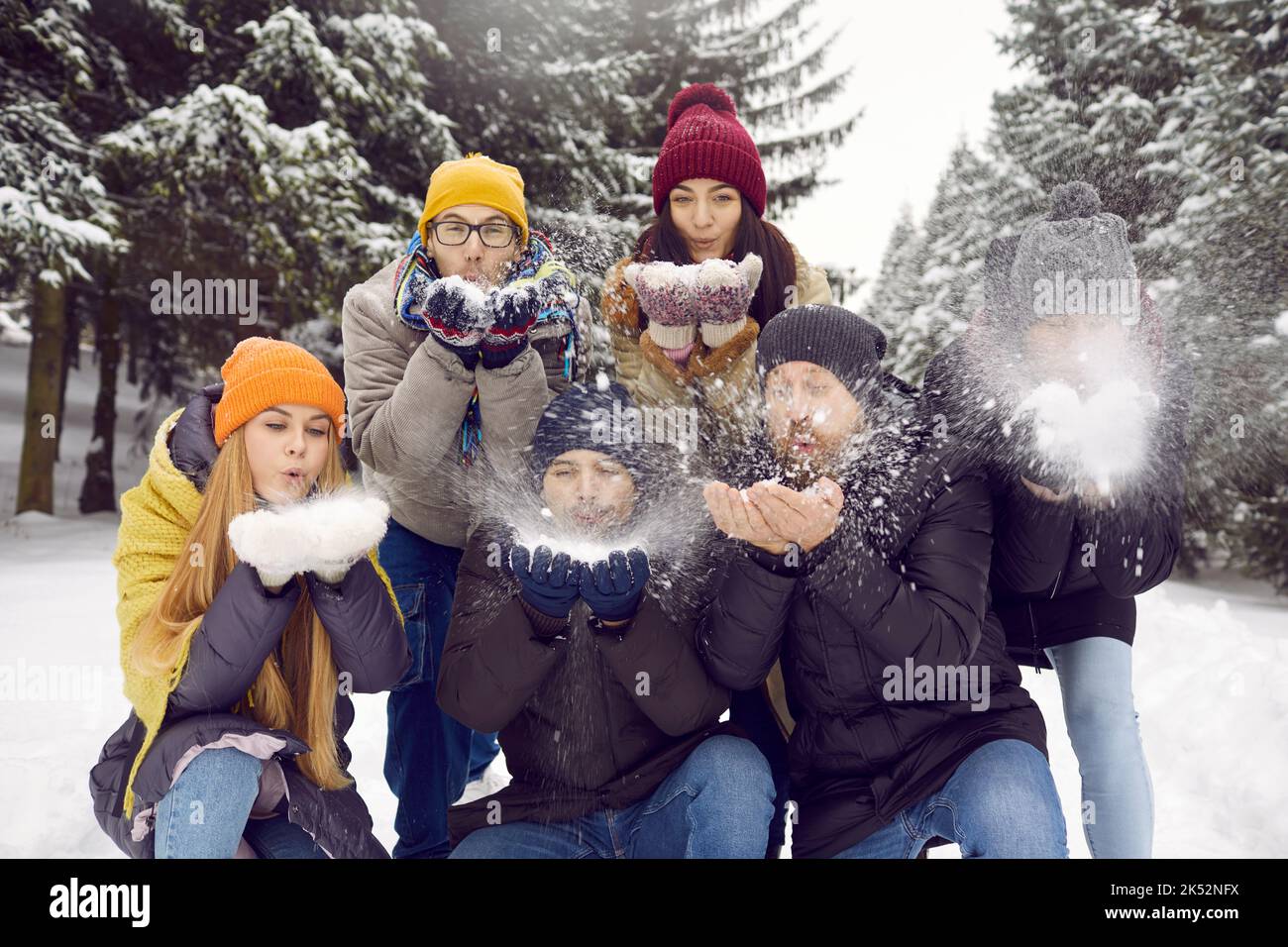 Eine Gruppe junger Freunde, die Schnee von den Händen blasen, während sie sich im verschneiten Wald oder Park Vergnügen. Stockfoto