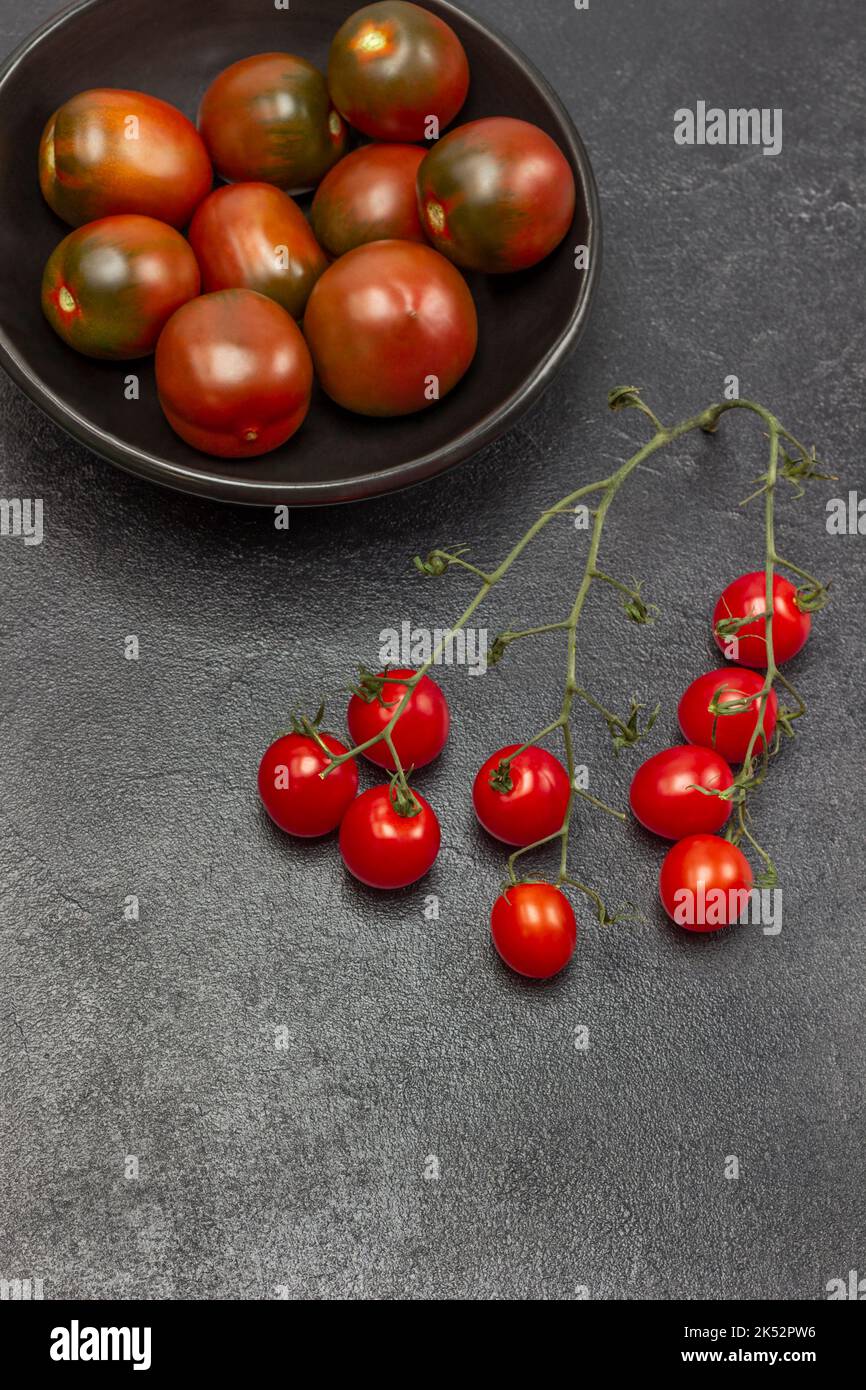 Zweig aus Kirschtomaten, rote Tomaten in schwarzer Schüssel. Speicherplatz kopieren. Flach liegend. Schwarzer Hintergrund. Stockfoto