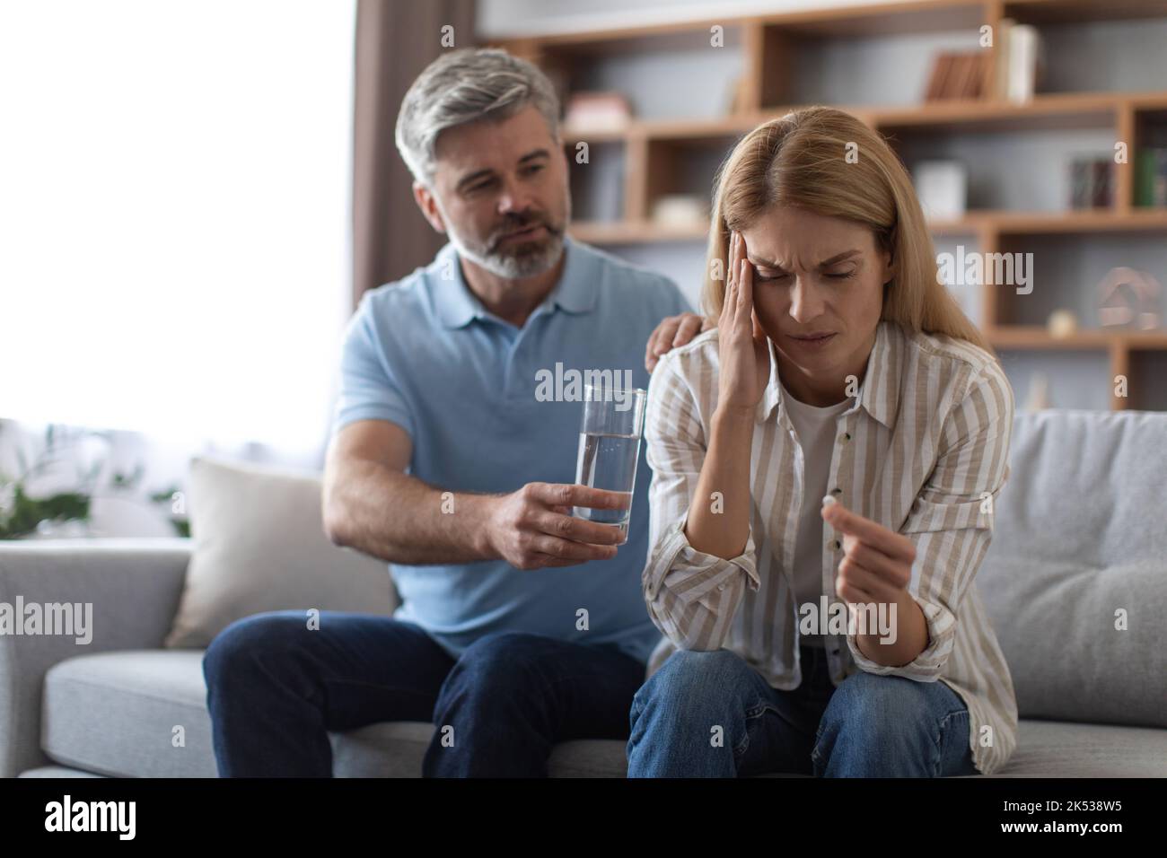 Besorgniserregender, reifer kaukasischer Ehemann beruhigt seine Frau, leidet unter Kopfschmerzen und gibt ein Glas Wasser auf das Sofa Stockfoto