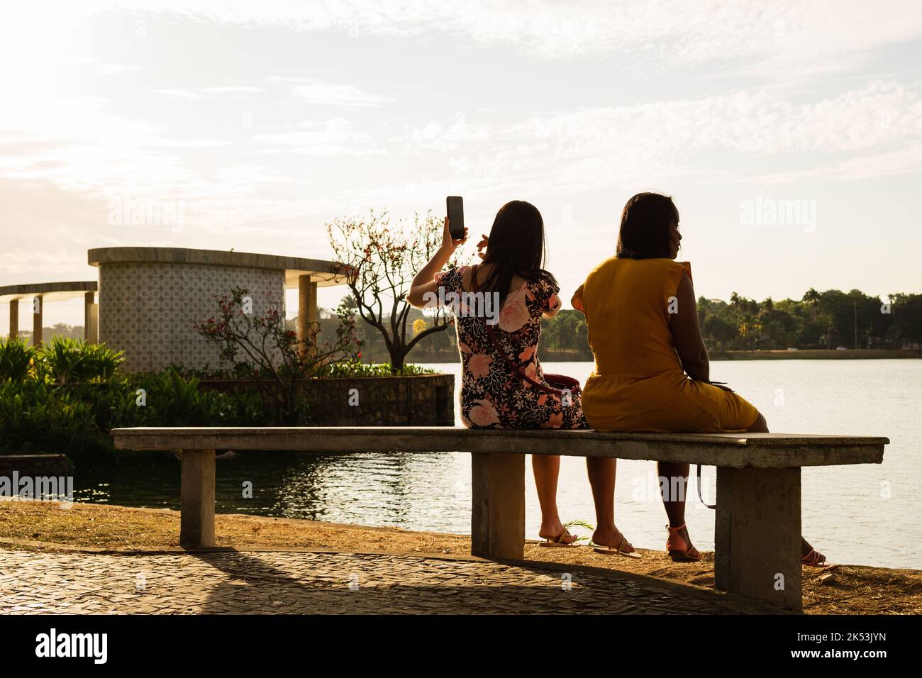 Zwei Frauen saßen auf einer Bank und nahmen ein Foto mit einem Handy der Casa do Baile in Lagoa da Pampulha in Belo Horizonte, Minas Gerais, Brasilien, auf. Stockfoto