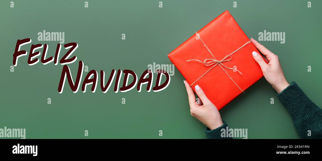 Weibliche Hände mit Geschenk und Text FELIZ NAVIDAD (Spanisch für Frohe Weihnachten) auf grünem Hintergrund, Draufsicht Stockfoto
