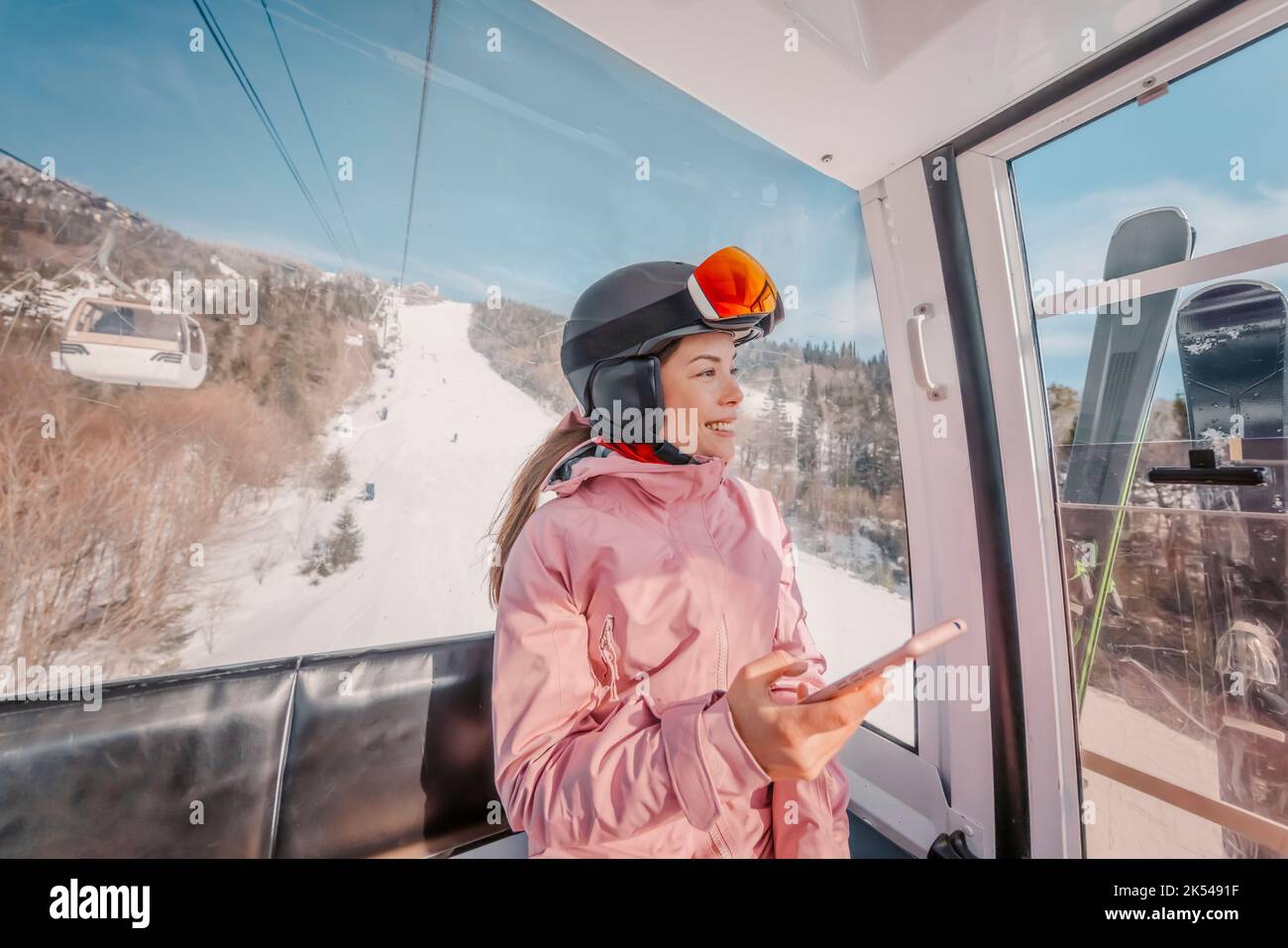 Skiurlaub - Frau Skifahrerin mit Telefon-App in Gondelbahn. Lächelndes Mädchen mit dem mobilen Smartphone, das Skibekleidung, Helm und Brille trägt. Ski Stockfoto