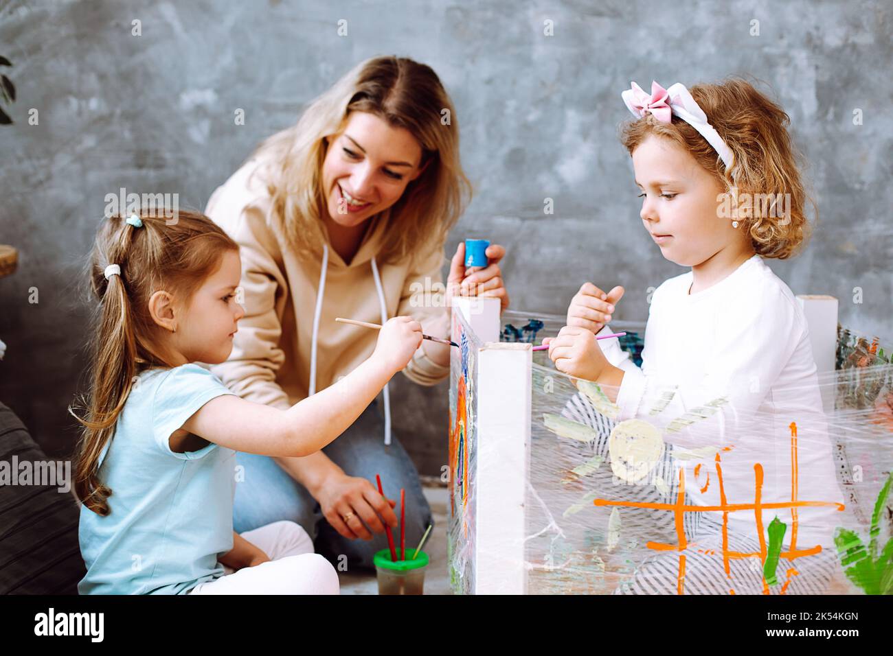 Nette Erzieherin helfen kleinen Mädchen künstlerisches Handwerk im Spielzimmer des Kindergartens zu machen. Kinder sitzen und zeichnen mit leuchtenden Farben und Pinsel auf dem Gestell Stockfoto