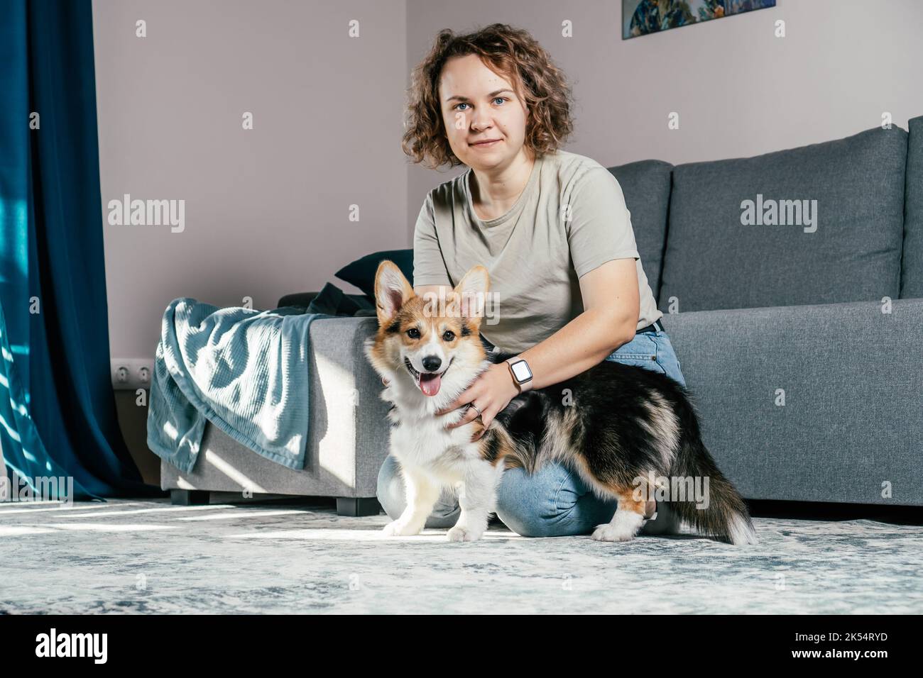 Lockig behaarte, fröhliche Barfußfrau mit Hundecorgi, die auf dem Boden sitzt, sich entspannt und im komfortablen Wohnzimmer neben dem Sofa spielt. Aktiv und Stockfoto