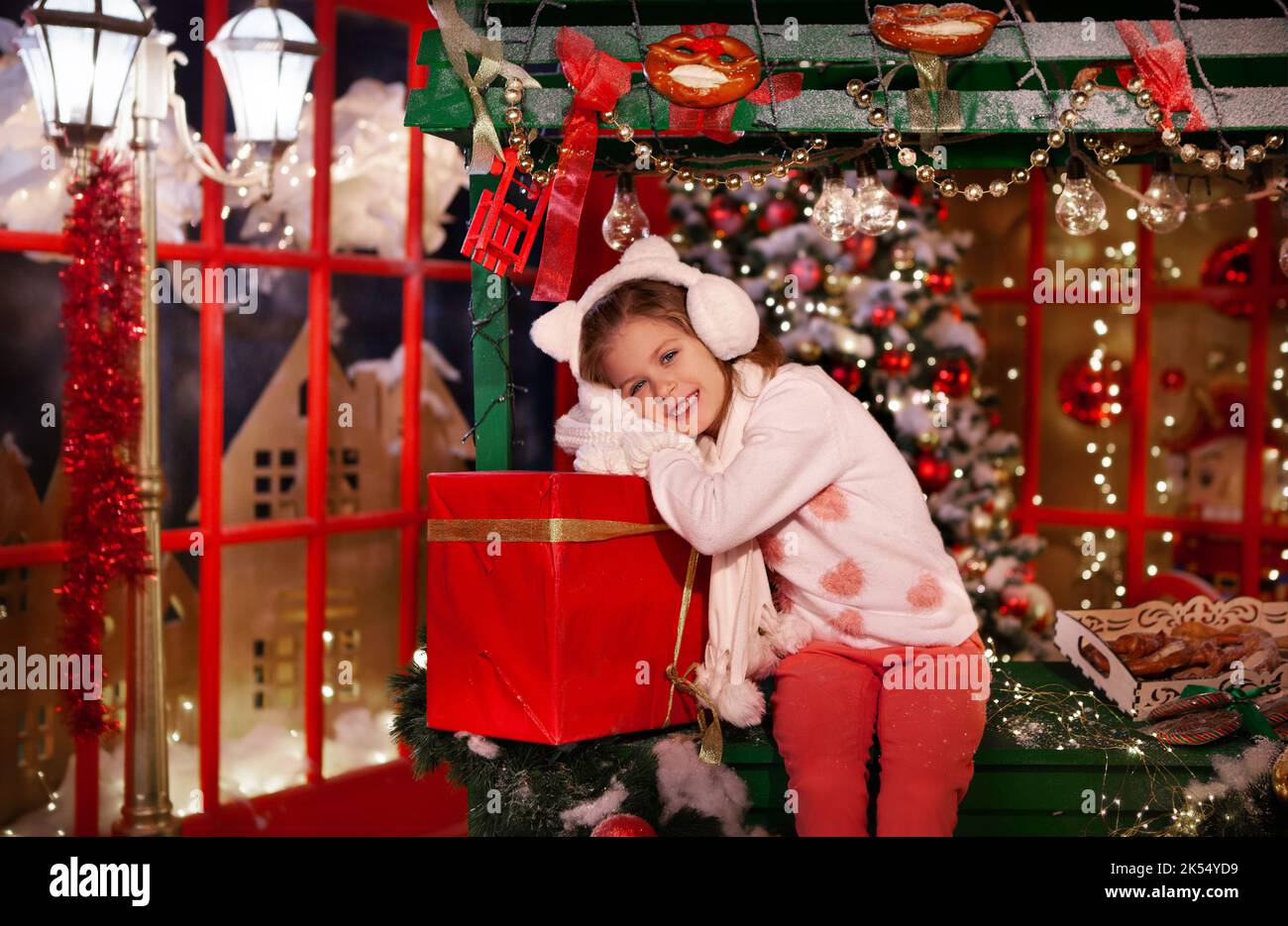 Little Pretty Girl hält rote Geschenkbox mit Band. Model lächelt, schaut auf die Kamera und trägt Pullover, Fäustlinge, Schal und Katzenohren Stockfoto