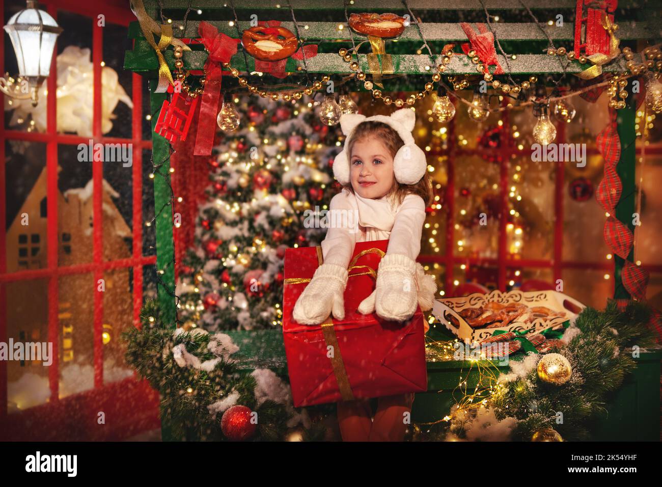 Schönes Mädchen überrascht von Weihnachtsgeschenk. Niedliches Modell mit Pullover, Fäustlingen und Katzenohren. Kind auf dem Hintergrund des roten Schaufensters an der Christm Stockfoto