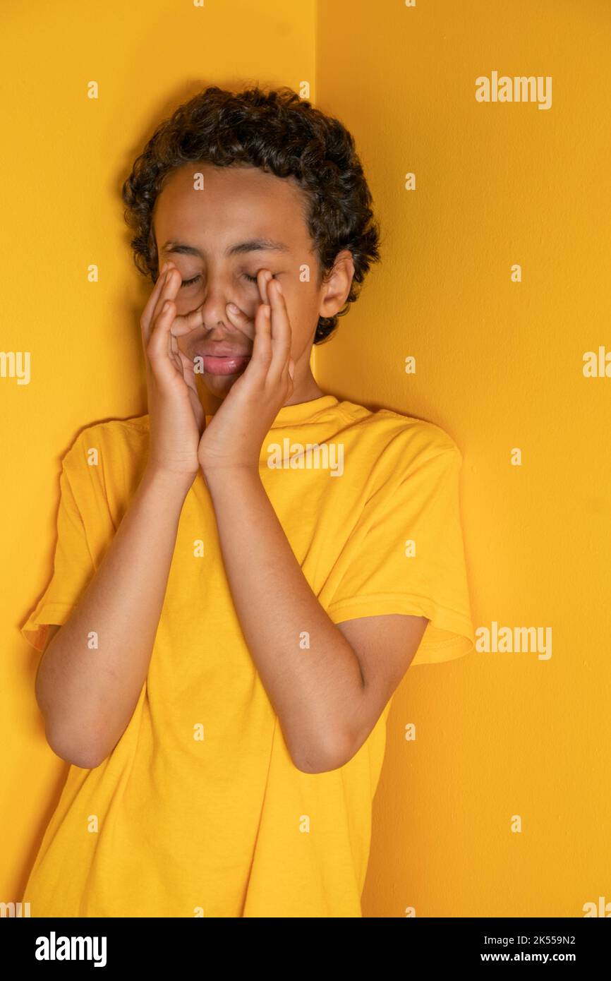 Ethnischer 12-jähriger Junge mit einem gelben/orangen Hemd, das niesen kann, an der gleichen gelb/orangen Wand Stockfoto