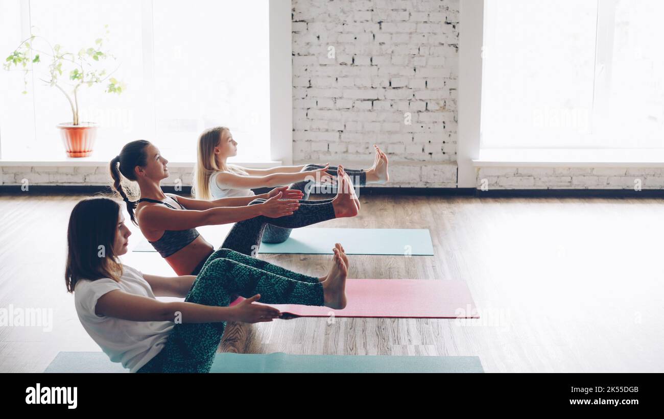 Pan Shot von schlanken jungen Damen, die Yoga üben und dabei eine Balancierbootpose in einem hellen, geräumigen Raum machen. Junge Frauen konzentrieren sich auf Gegengewicht, sie üben gerne. Stockfoto