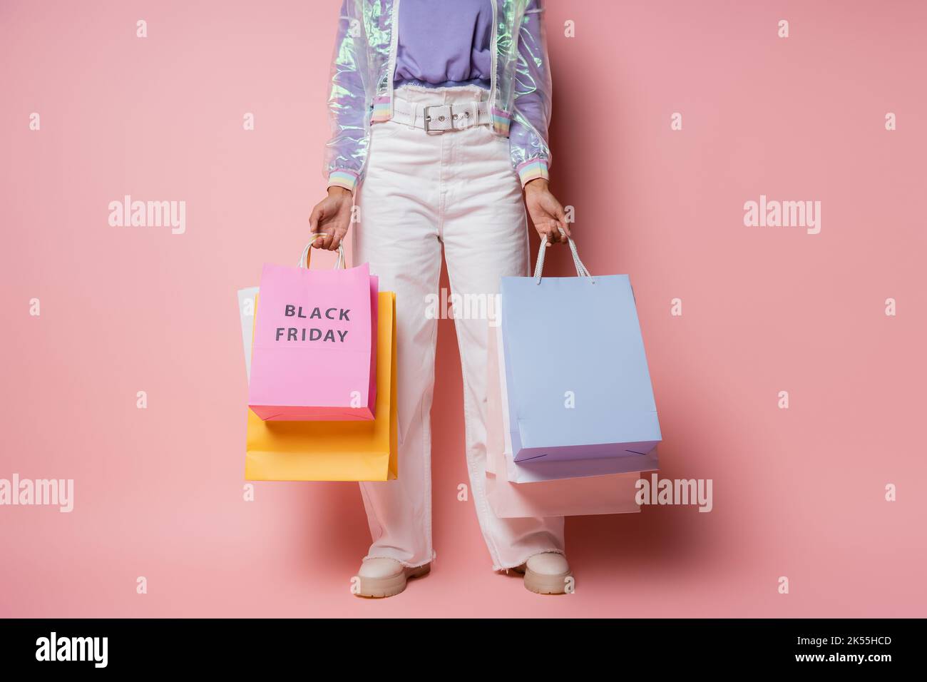 Beschnittene Ansicht der Frau in transparenter Jacke und weißer Hose mit Einkaufstaschen mit schwarzem freitag-Schriftzug auf rosa, Stockbild Stockfoto