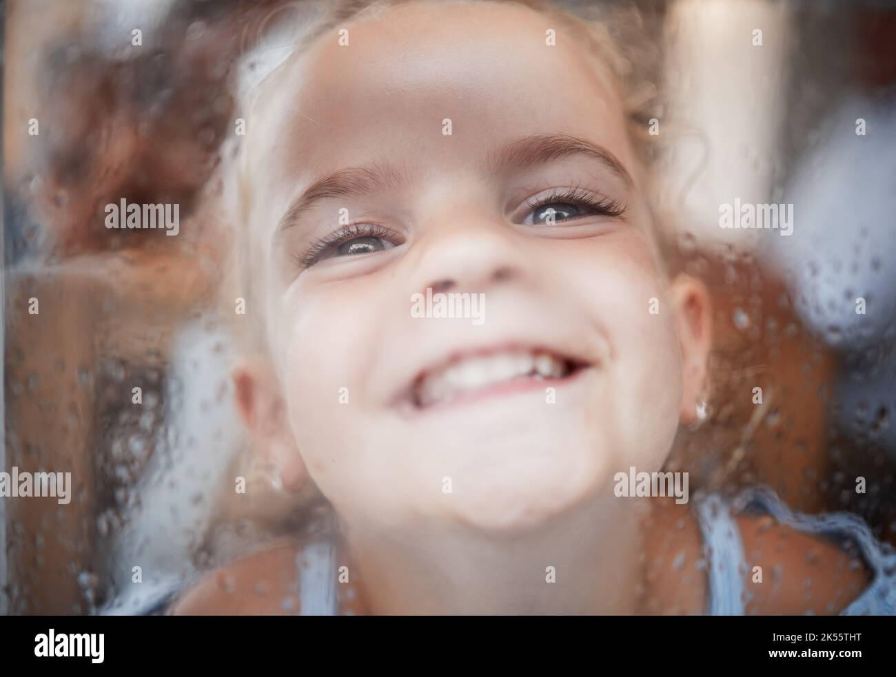 Fröhliches lustiges Mädchen, Gesicht am Fenster mit flacher Nase auf regnerischem Glas und Comic-Porträt zu Hause. Junges Kind spielen Witz, Kind allein mit großem Lächeln und Stockfoto