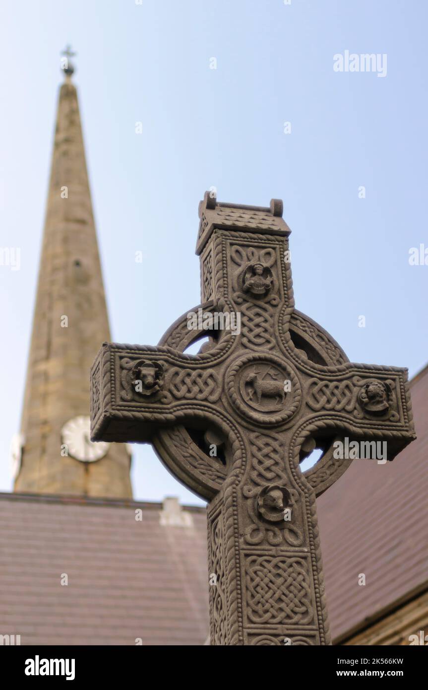 Kunstvoll geschnitztes keltisches Kreuz mit Knoten und dahinter der Kirchturm. Stockfoto