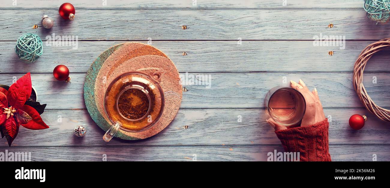 Wintertee, Panorama-Flachlage mit Glas-Teekannen, Glas Tee in der Hand auf verblassten Holzplanken. Weihnachtsdekor - Spiegelkugeln, Kugeln, Spielzeug Stockfoto