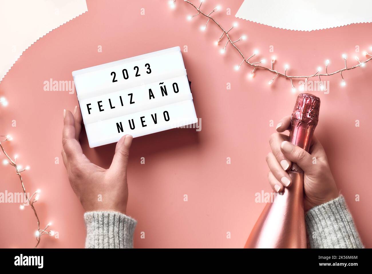 Text Frohes neues Jahr 2023 in spanischer Sprache auf Leuchtkasten. Leichte Girlande, Frauenhänden mit Leuchtkasten und pinker Champagner-Flasche in Metallic-Optik. Neujahr Stockfoto