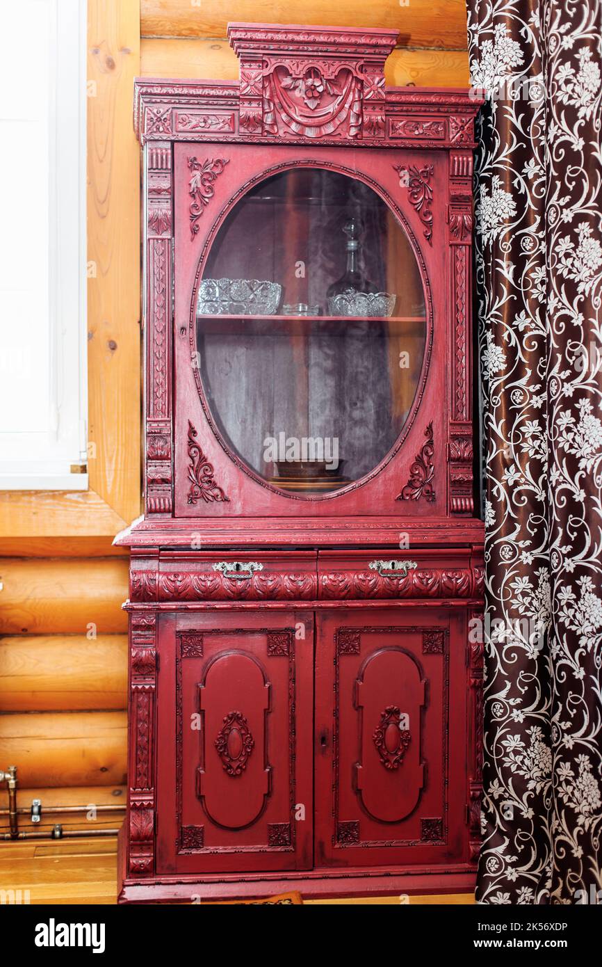 Alter alter, roter Holzschrank mit geschnitzten Dekorationen in der Ecke einer ländlichen Blockhütte zwischen Fenster und braunem Vorhang. Stockfoto