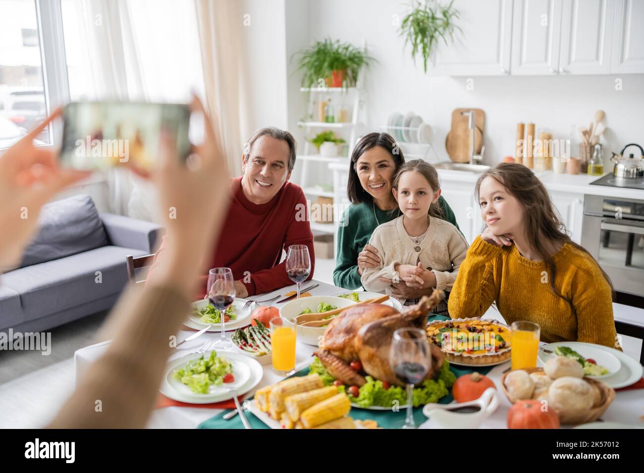 Multiethnische Familie, die in der Nähe des Danksagesessens sitzt, während die Frau zu Hause auf dem Smartphone fotografiert, Stockbild Stockfoto