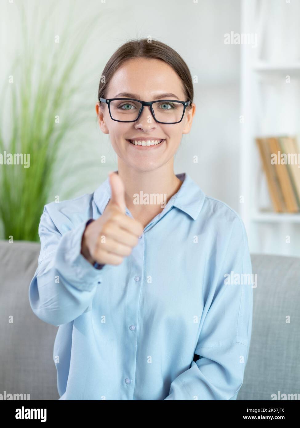 Erfolgreiche Frau. Wie Geste. Gute Arbeit. Glückwunsch zum Erfolg. Zufriedene, stolz lächelnde, intelligente Trainerin, die mit dem Daumen nach oben auf der Couch steht Stockfoto
