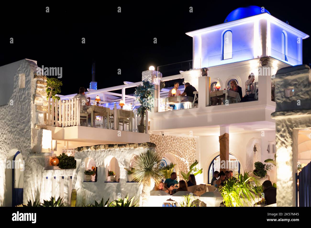 Eine griechische Taverne, in der Gäste an einem warmen Sommerabend an Tischen im Freien speisen. Das Restaurant ist mit blau-weißer Beleuchtung beleuchtet Stockfoto