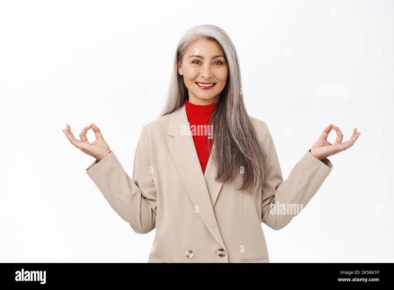 Behaltet die Ruhe. Asiatische ältere Frau im Business-Anzug, meditierend, tief durchatmen, Luft einatmen und sich entspannen, stehend über weißem Hintergrund Stockfoto