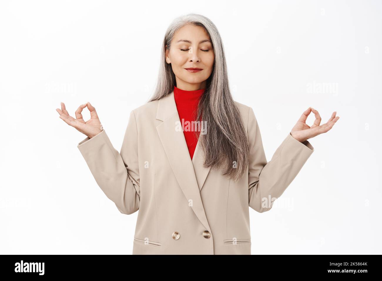 Behaltet die Ruhe. Asiatische ältere Frau im Business-Anzug, meditierend, tief durchatmen, Luft einatmen und sich entspannen, stehend über weißem Hintergrund Stockfoto