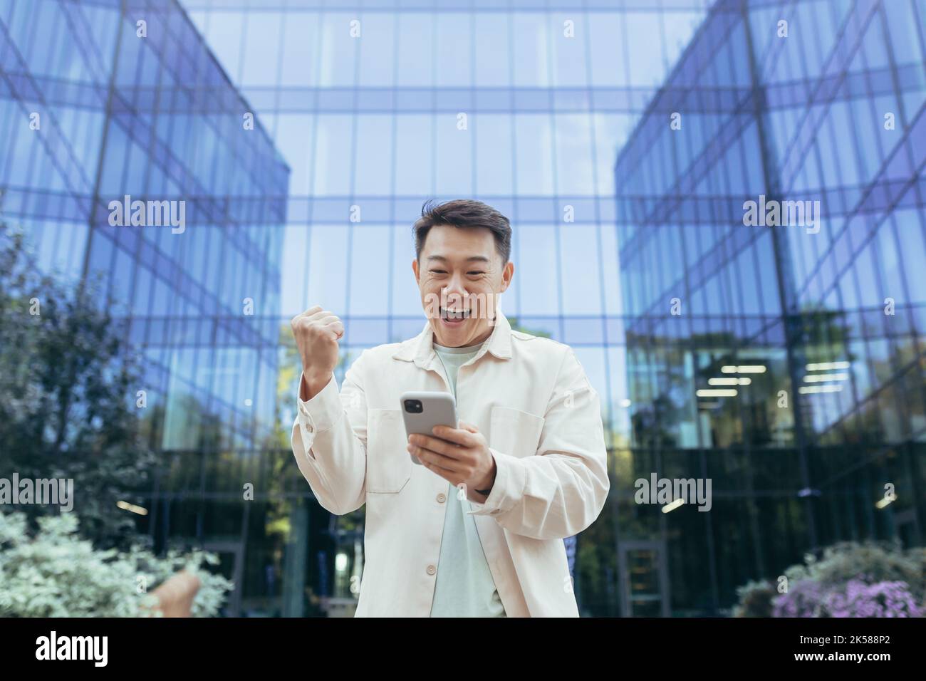 Glücklicher asiatischer Mann, der Sieg und Erfolg feiert, das Telefon lächelnd und glücklich hält, Programmierer außerhalb des modernen Bürogebäude-Softwareunternehmens, Mann in legerer Kleidung mit Smartphone. Stockfoto