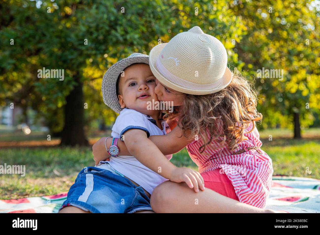 Zwei kleine Geschwister sitzen auf einer Picknickdecke im Park, Schwester küsst den kleinen Bruder auf die Wange. Stockfoto