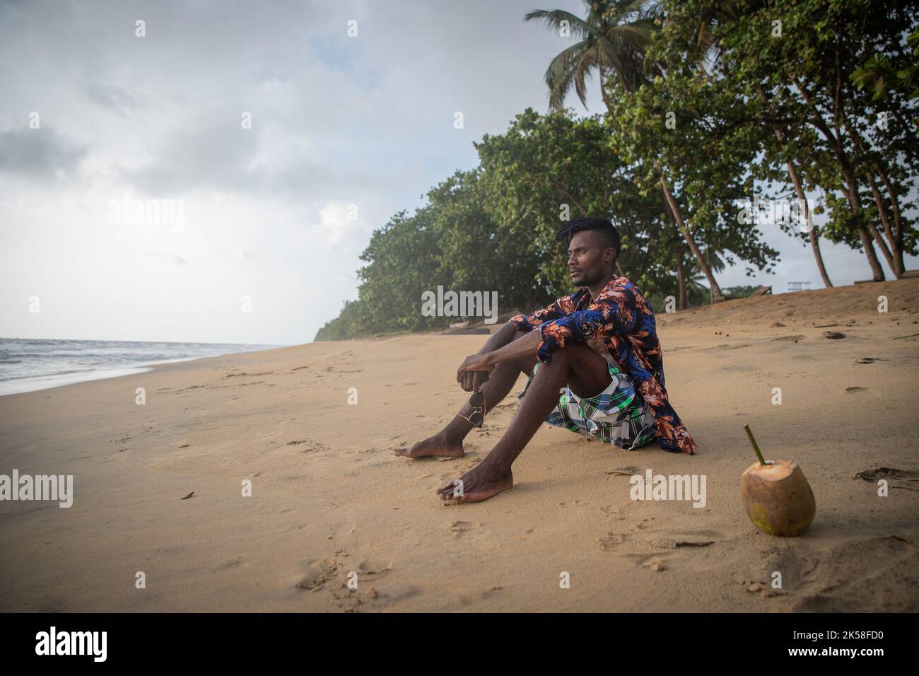 Junger Mann sitzt pensiver auf dem Strandsand, hält seine Sonnenbrille und genießt sein Leben, Kopierer Platz auf der linken Seite. Stockfoto