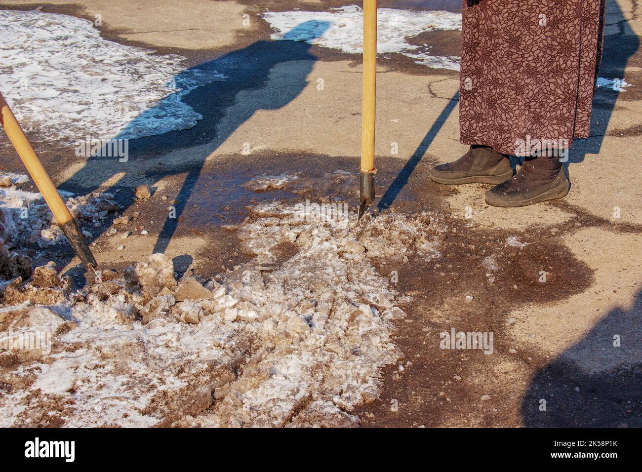 Eine Arbeiterin reinigt das Eis und entfernt mit einem Eisbrecher Schnee von den Pflasterplatten. Ein Mann bricht Eis mit einem Stahlklingenbrecher, ein Eisbrecher ebenfalls Stockfoto