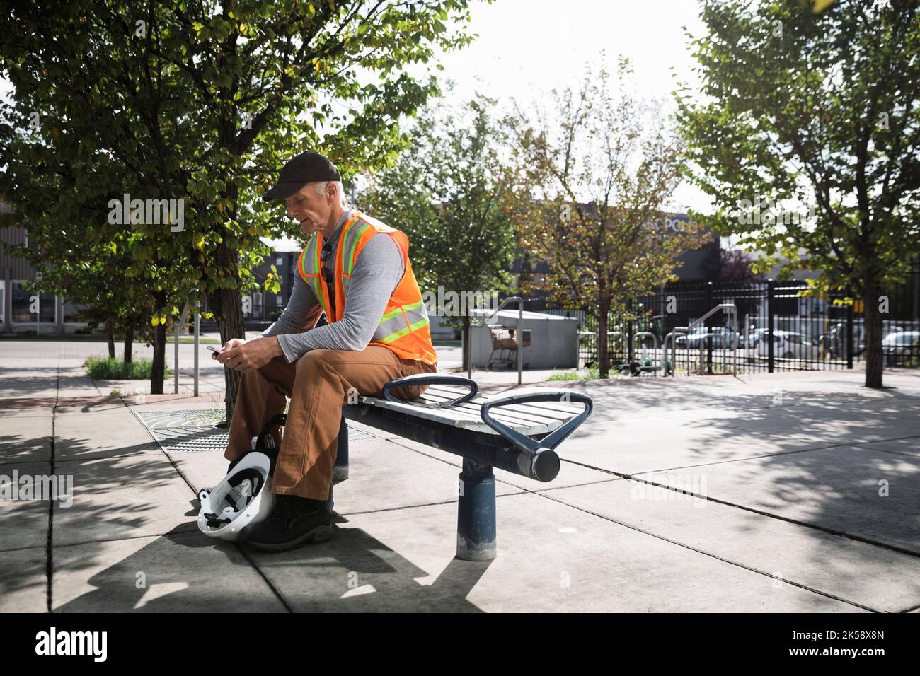 Leitender Arbeiter in hoher Visusjacke, der mit dem Smartphone auf der Bank sitzt Stockfoto