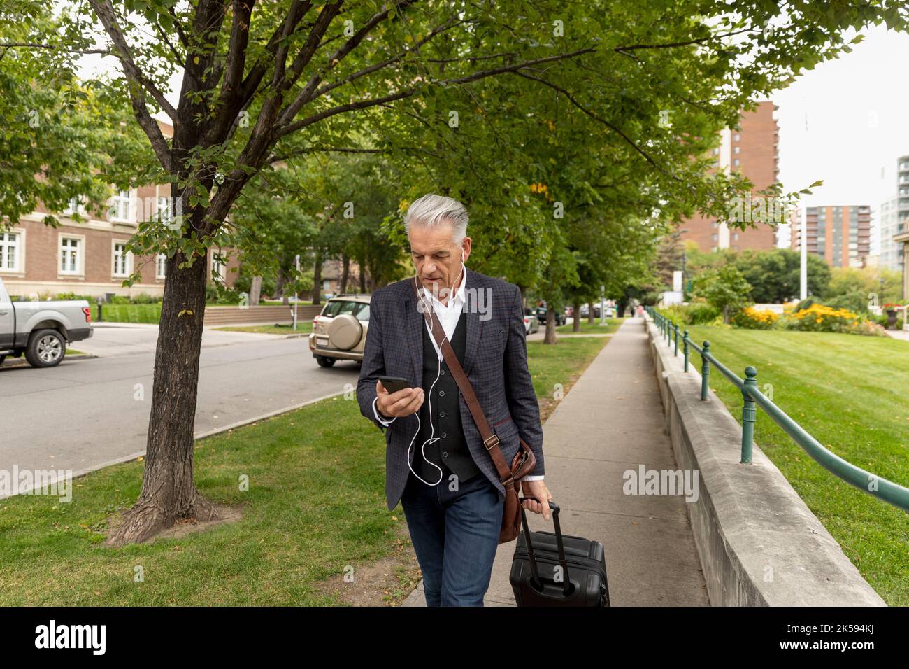 Leitender Geschäftsmann mit Kopfhörern und Smartphone, der den Koffer auf dem städtischen Bürgersteig zieht Stockfoto