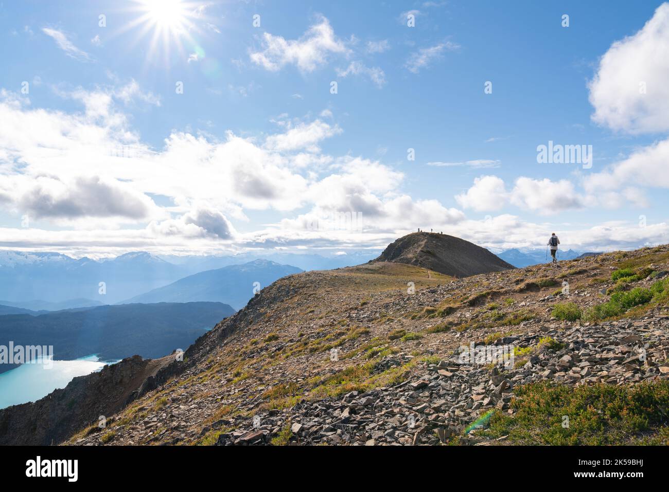 Eine Frau wandert entlang des Panorama Ridge mit dem Garibaldi Lake weit unten und dem Coastal Mountain Range und strahlender Sonne in der Ferne. Stockfoto