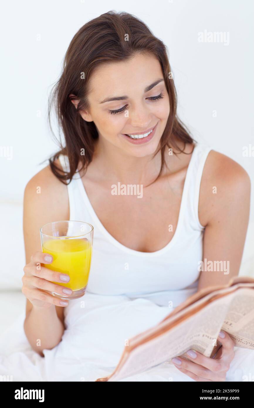 Mein morgendliches Ritual. Eine junge Frau liest die Zeitung im Bett mit einem Glas Orangensaft in der Hand. Stockfoto