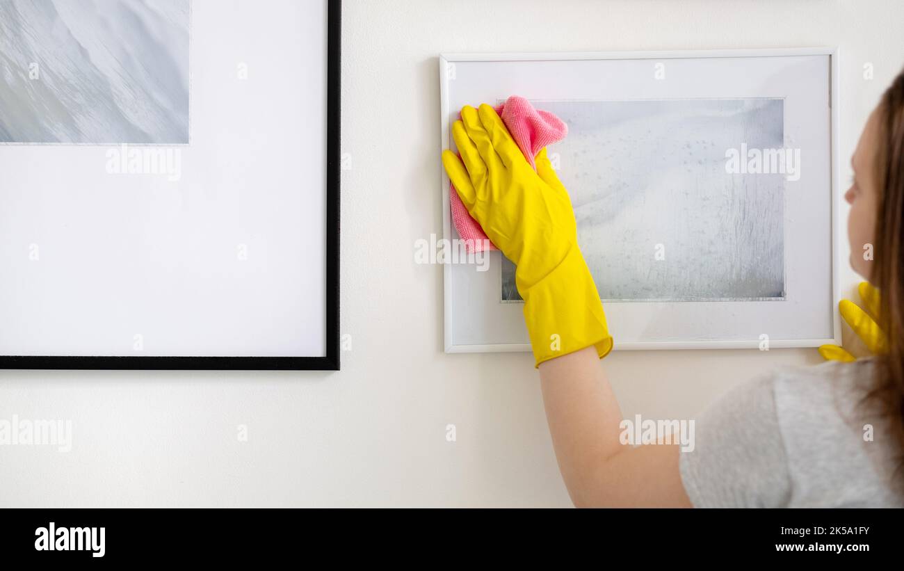 Hausreinigung. Hausmeisterin. Hausarbeit Routine. Nicht erkennbare Frau in gelben Schutzhandschuhen, die Bild-Kunst von Staub-Mikrofasertuch entfernen. Stockfoto