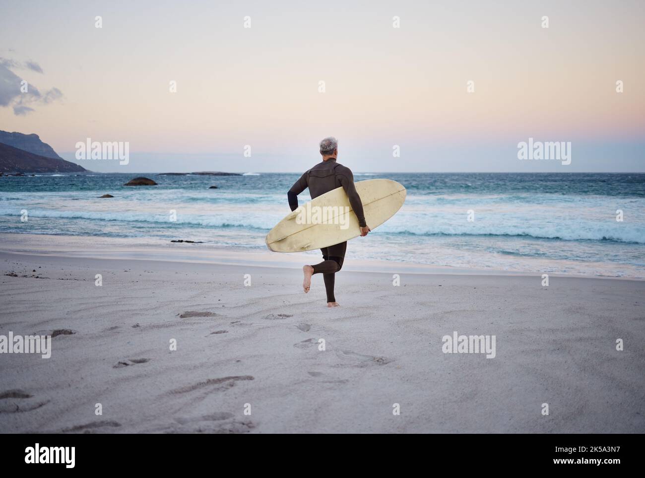Surfen Sie am Strand, Mann und laufen zum Meer mit Surfbrett für die Wellen, Fitness und Abenteuer. Reifes Surfer bei Sonnenaufgang, im Nassanzug und zum Genießen Stockfoto