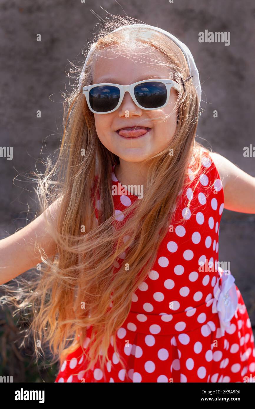 Schönes glückliches kleines Mädchen in einem roten Kleid mit Punktmuster, das lächelnd Zunge auf Betonwand-Hintergrund zeigt. Nettes fröhliches Kind mit langen blonden Haaren in su Stockfoto
