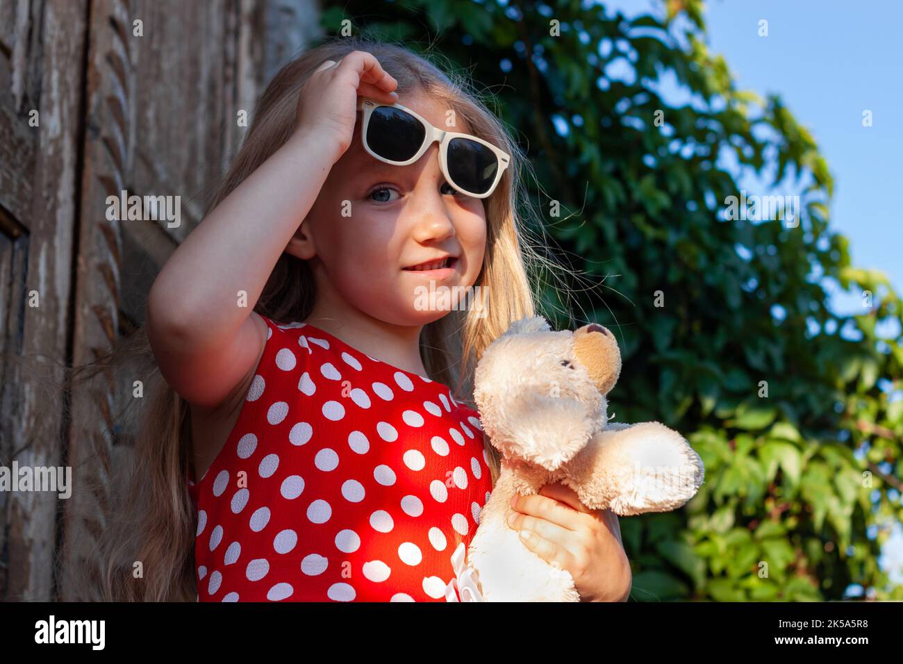 Schöne glückliche Mädchen in roten Polka dot Kleid mit Hund Plüsch-Spielzeug lächelnd auf Holzbalkon. Nettes fröhliches Kind mit langen blonden Haaren in Sonnenbrillen auf gree Stockfoto