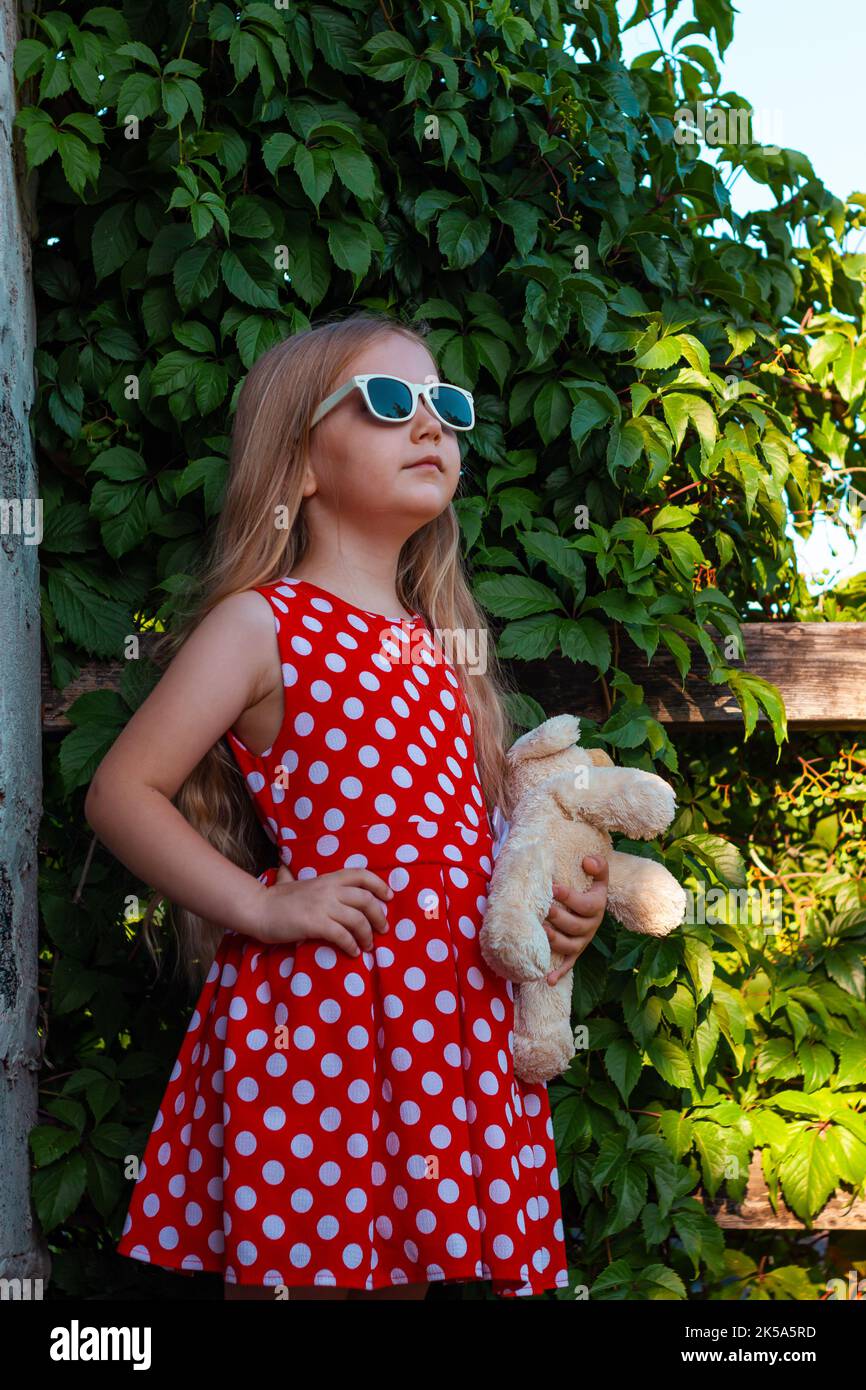 Schöne glückliche Mädchen in roten Polka dot Kleid mit Hund Plüsch-Spielzeug lächelnd auf Holzbalkon. Nettes fröhliches Kind mit langen blonden Haaren in Sonnenbrillen auf gree Stockfoto