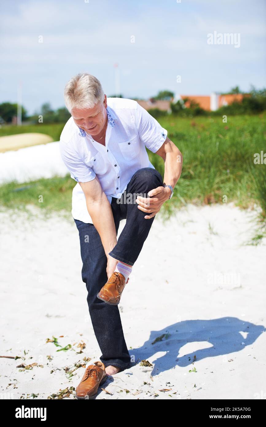 Er will den Strand unter seinen Füßen spüren. Ein älterer Mann, der seine Schuhe am Strand auszieht. Stockfoto