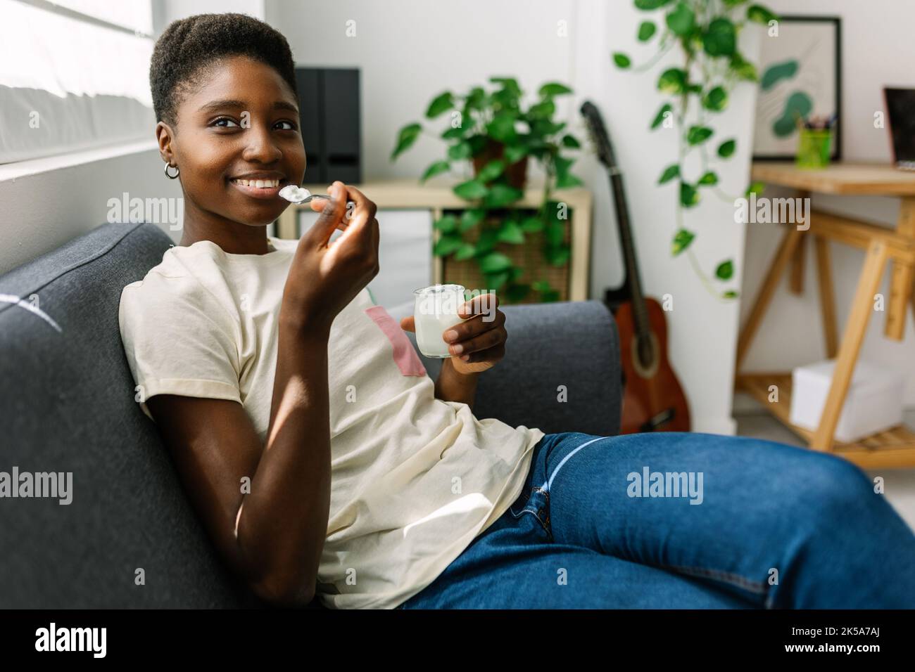 Glückliche junge afrikanische Frau, die sich auf dem Sofa entspannt und Bio-Joghurt isst Stockfoto