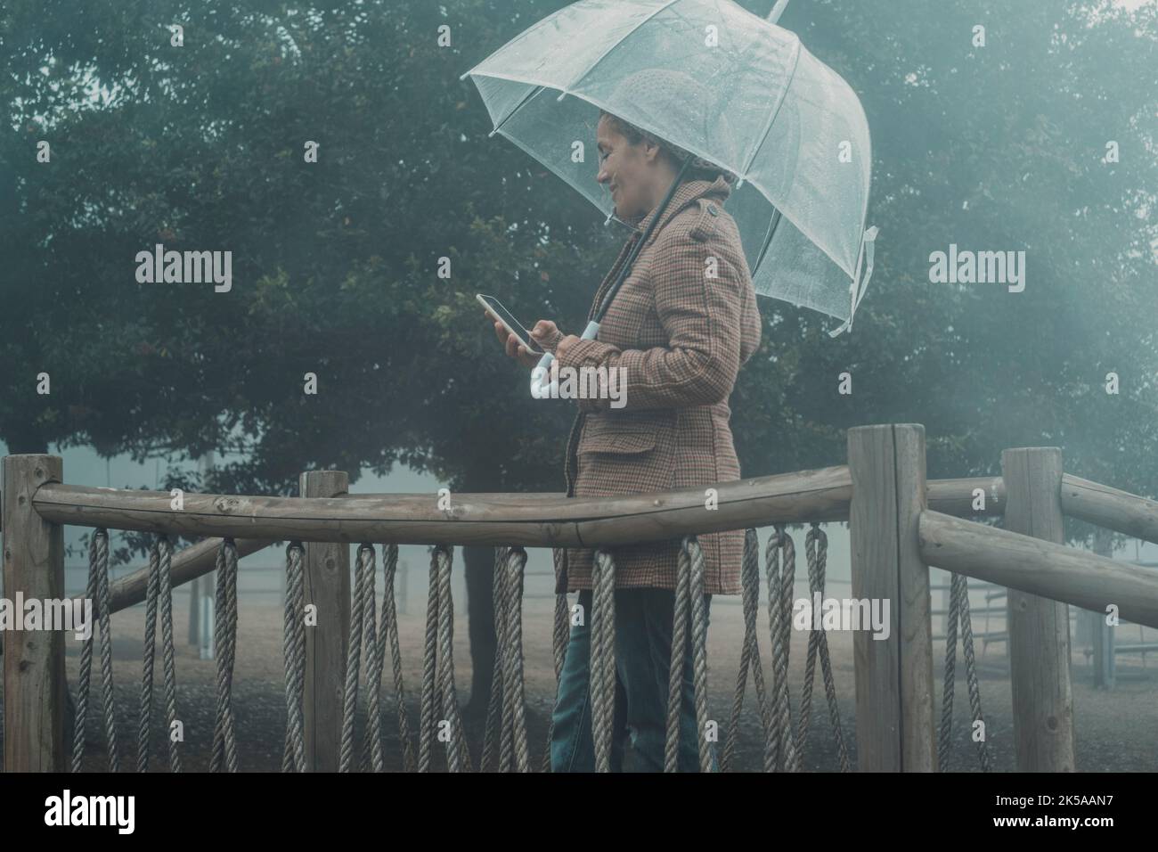 Die Frau benutzt das Mobiltelefon unter dem Regen, geschützt durch einen transparenten Regenschirm. Regnerischer Tag und schlechtes Wetter im Park. Menschen in der Freizeit im Freien Stockfoto