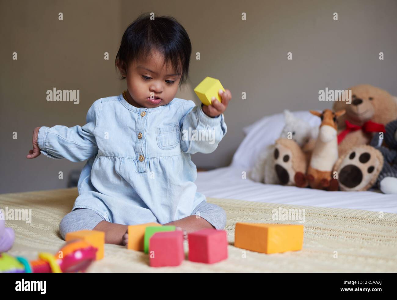 Down-Syndrom, Kindertagesstätte und Baby auf dem Bett mit Spielzeug, spielen mit Farbformen und Blöcken. Kinderentwicklung, besondere Bedürfnisse und nettes Mädchen im Schlafzimmer Stockfoto