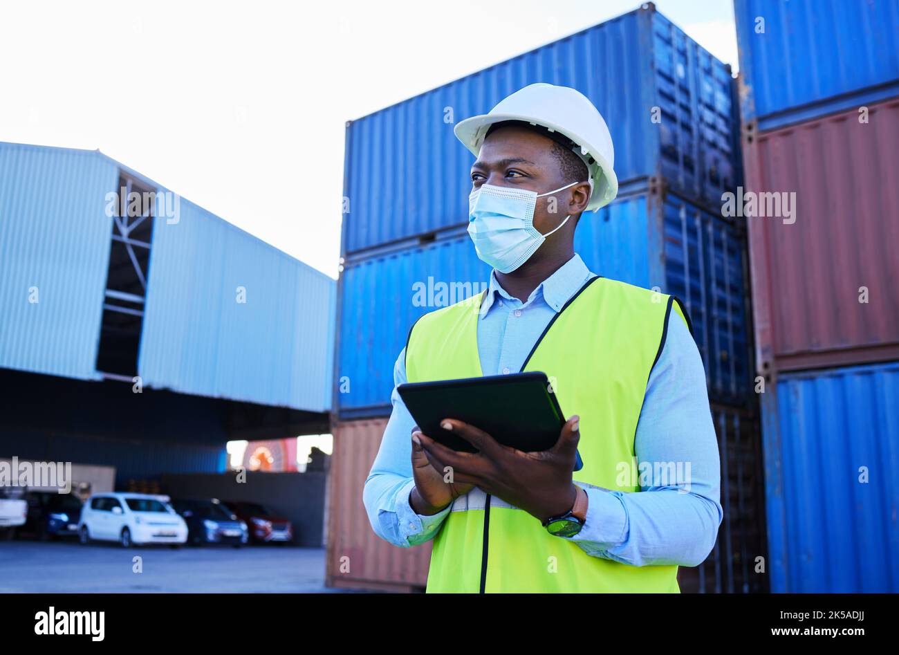 Mann, Logistikmitarbeiter und covid mit einem Tablet, um den Bestand in Maske mit Frachtcontainer im Hintergrund zu überprüfen. Mitarbeiter in der schwarzen Industrie arbeiten Stockfoto